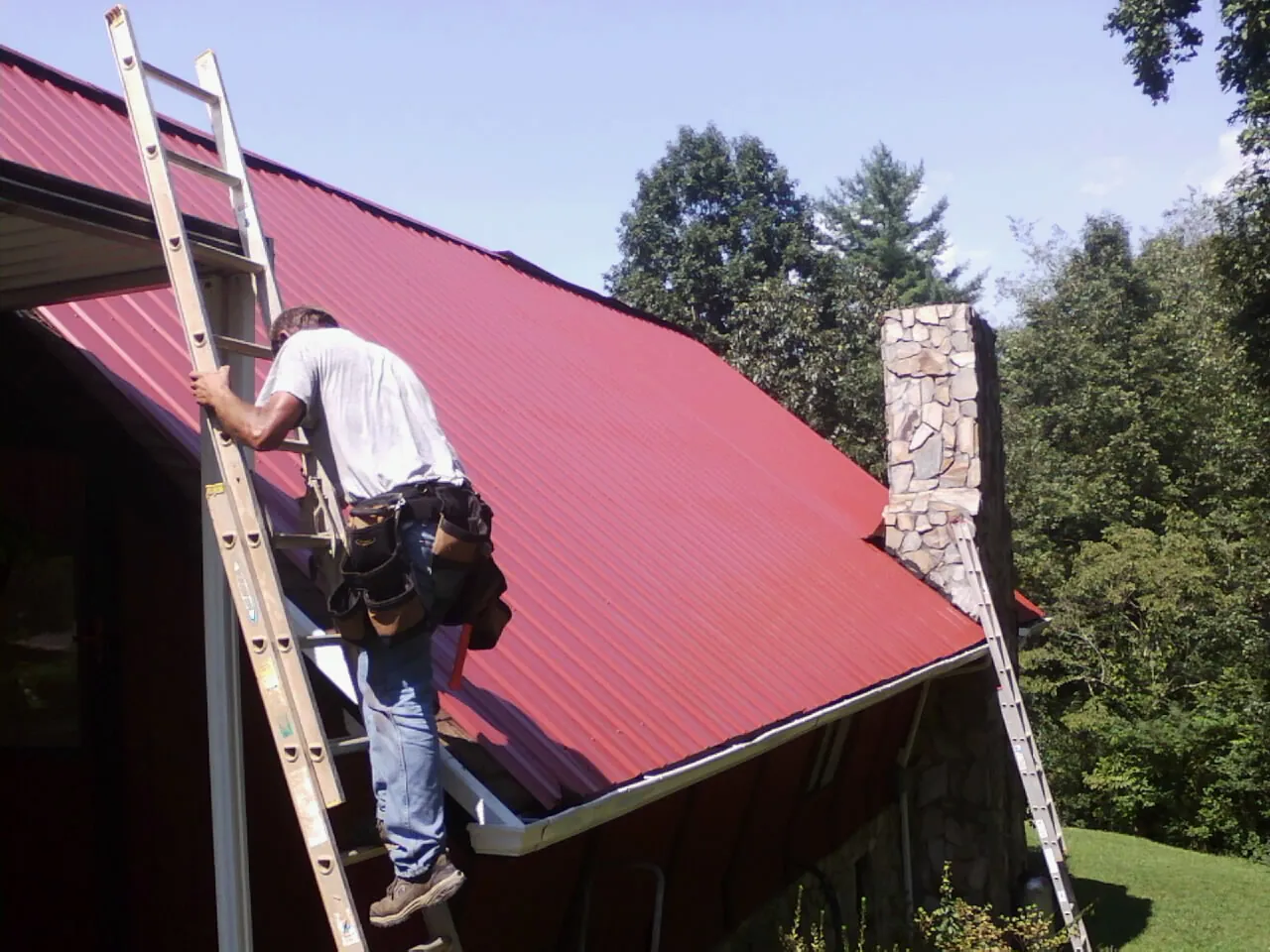 Roofer on a ladder beside a red metal roof and stone chimney — Soffit & Fascia Repair by Roofing Brothers in Dayton